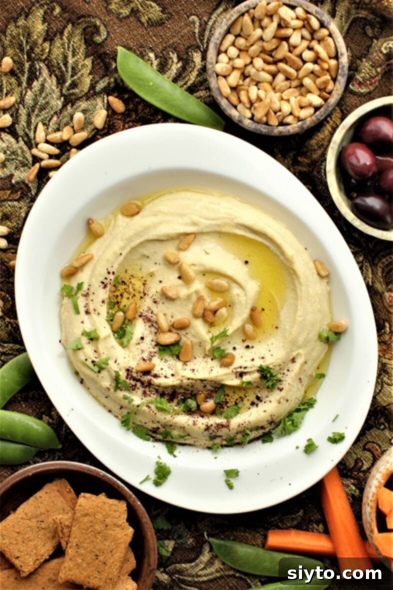A luscious bowl of ultra-creamy homemade hummus, elegantly garnished with a swirl of olive oil, toasted pine nuts, a dusting of vibrant sumac, and fresh parsley sprigs.