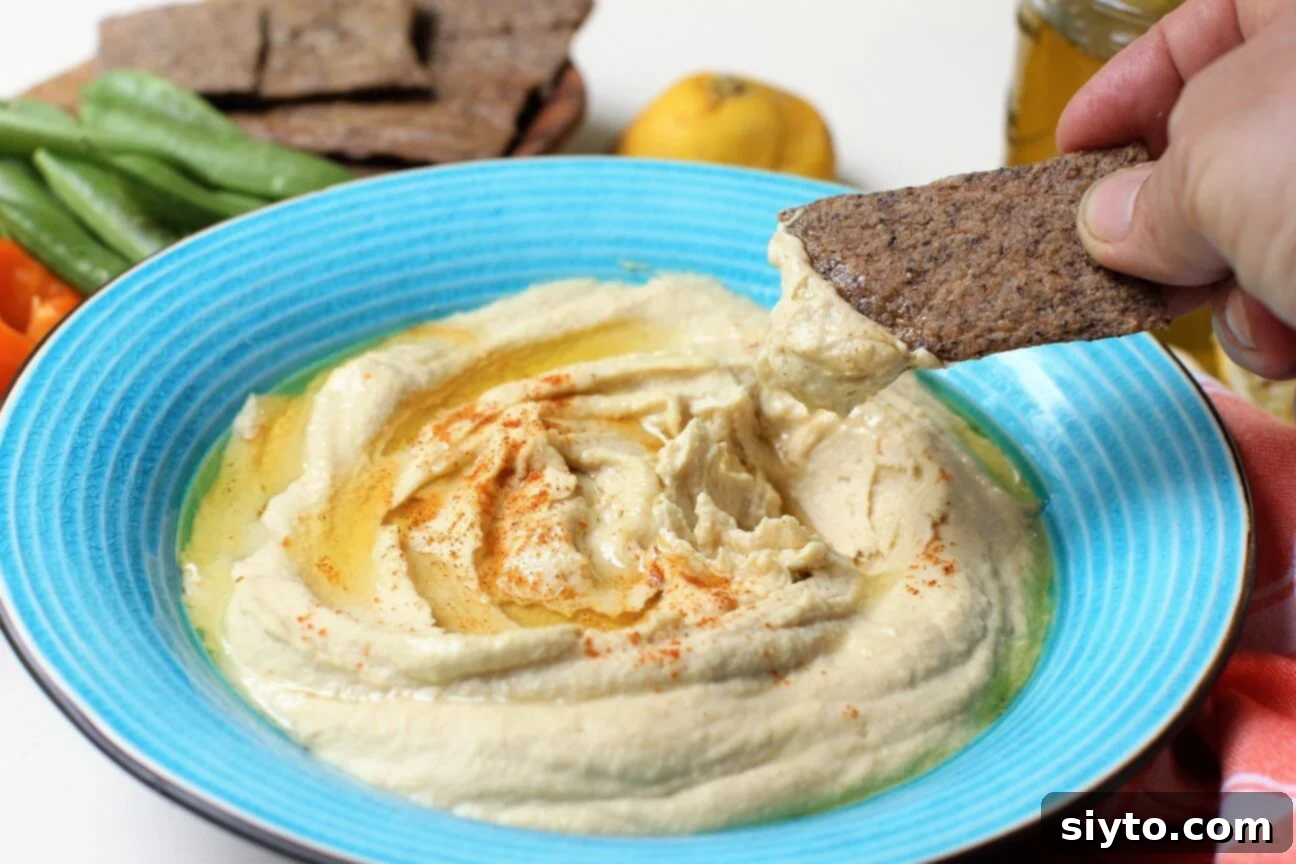 A hand dipping a crispy cracker into a bowl of creamy homemade hummus, highlighting its inviting and smooth texture, ready to be enjoyed.