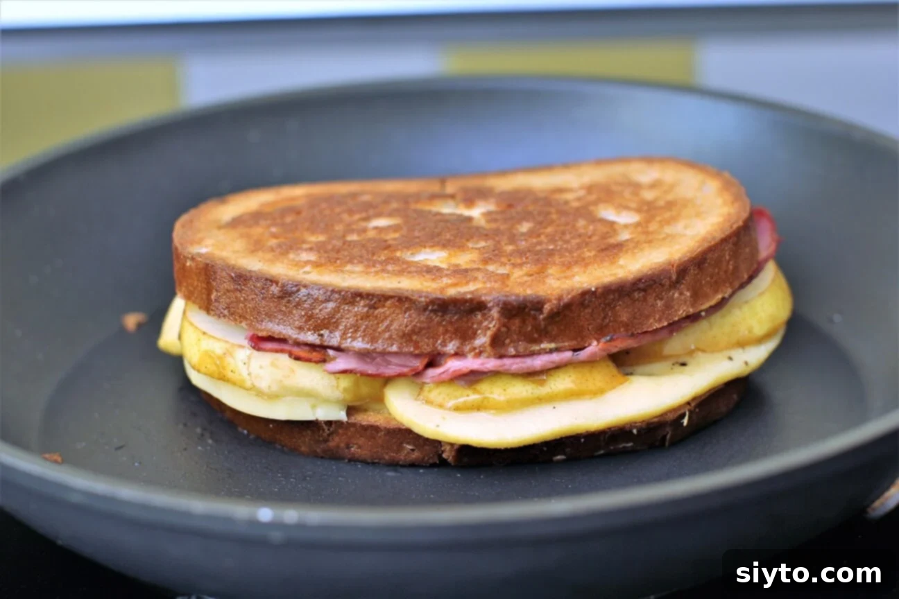 A pear and pastrami grilled cheese sandwich cooking in a hot skillet, becoming golden and crispy
