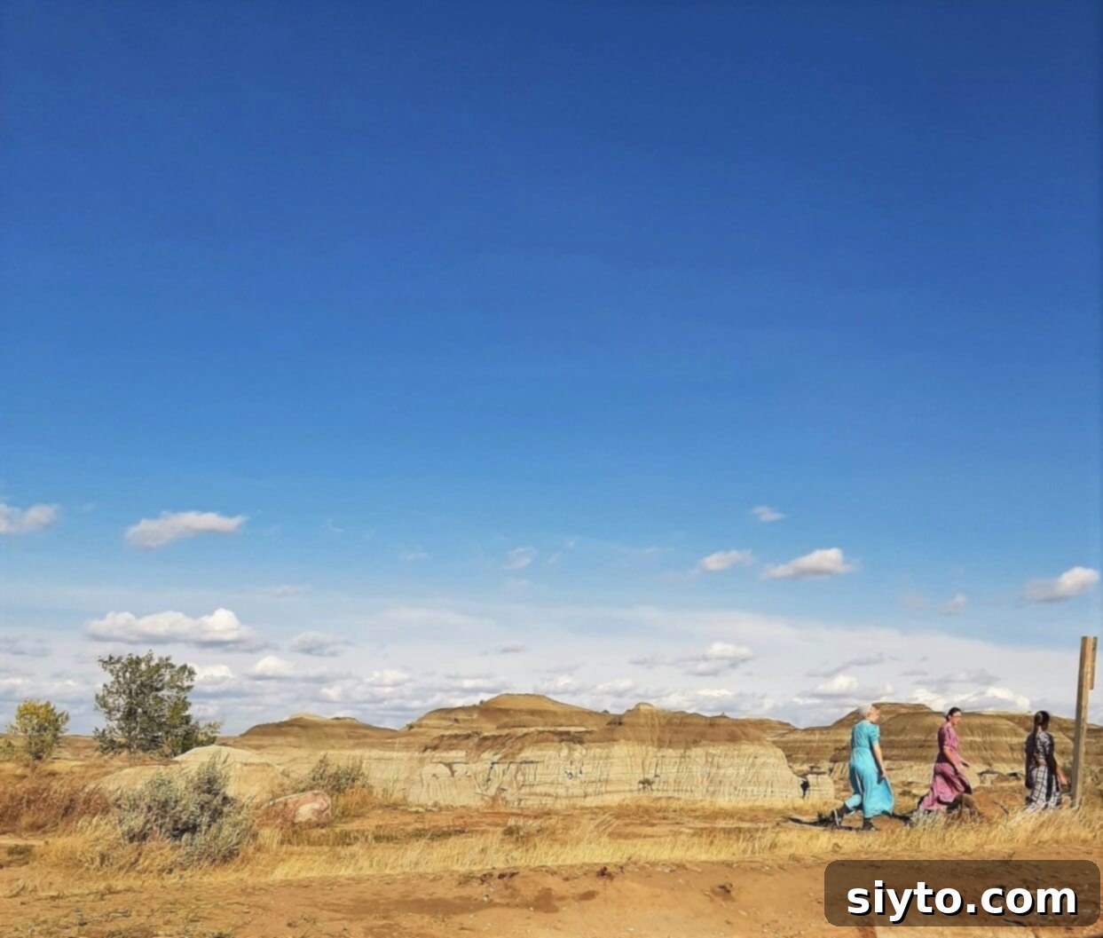 Three Mennonite girls in distinctive, bright dresses walking casually through the unique, arid landscape of Dinosaur Provincial Park.