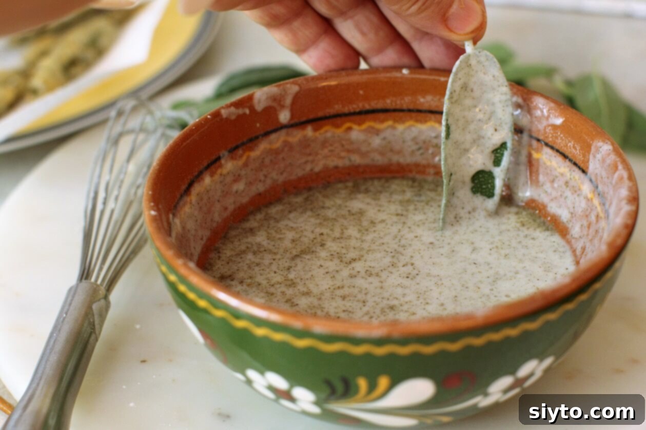A hand carefully dipping a fresh sage leaf into a light, creamy batter in a bowl, preparing it for frying.