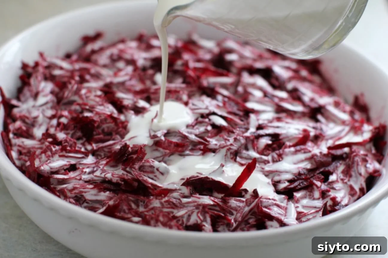 Close-up shot of heavy cream being slowly drizzled over a layer of fresh shredded beets and aromatics in a baking dish before cooking.