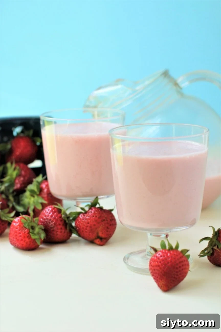 Two glasses of vibrant pink strawberry milk, garnished with fresh berries, sit next to a pitcher filled with the creamy drink, ready to be enjoyed on a hot day.