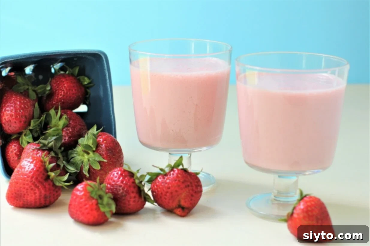 Two glasses of homemade strawberry milk, vibrant pink, are artfully arranged next to a scattering of fresh, whole strawberries on a wooden surface.