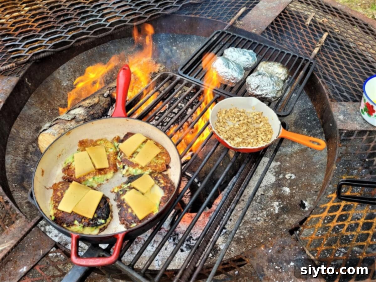 skillet of zucchini fritters bubbling with melting gouda cheese over a campfire, with oatmeal crumble topping nearby