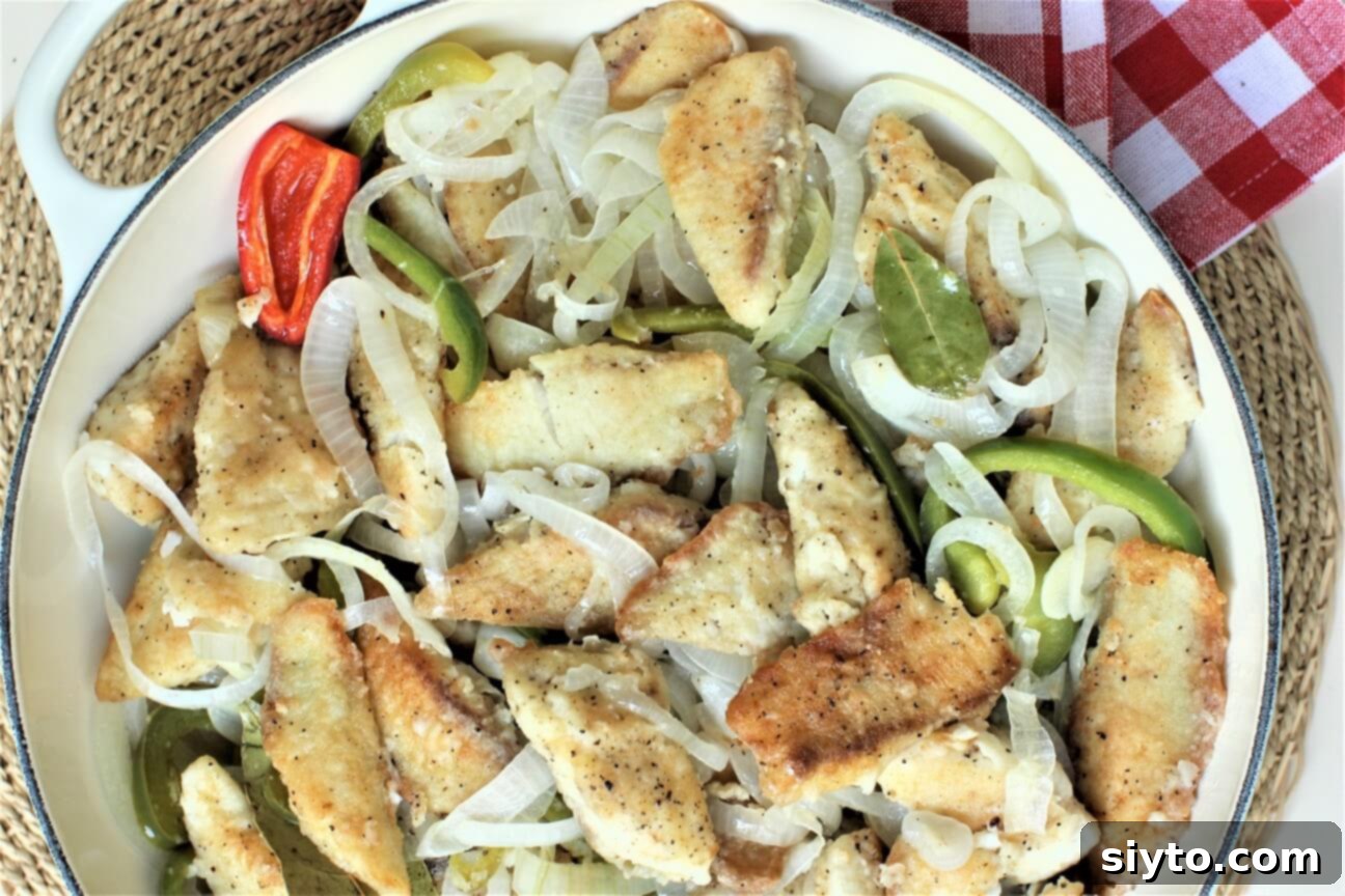 Top-down view of golden-fried tilapia fillets with onions and peppers, gently simmering in the tangy oil and vinegar sauce within a braiser.