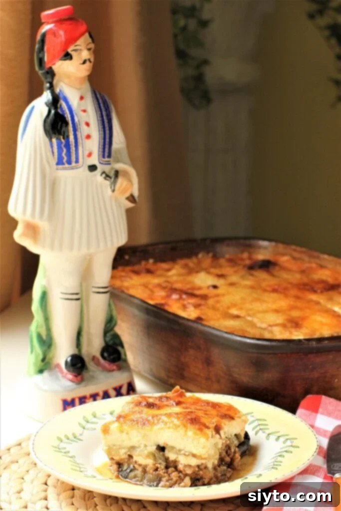 moussaka portion on plate, pan of moussaka behind, and statue of man in Greek traditional costume