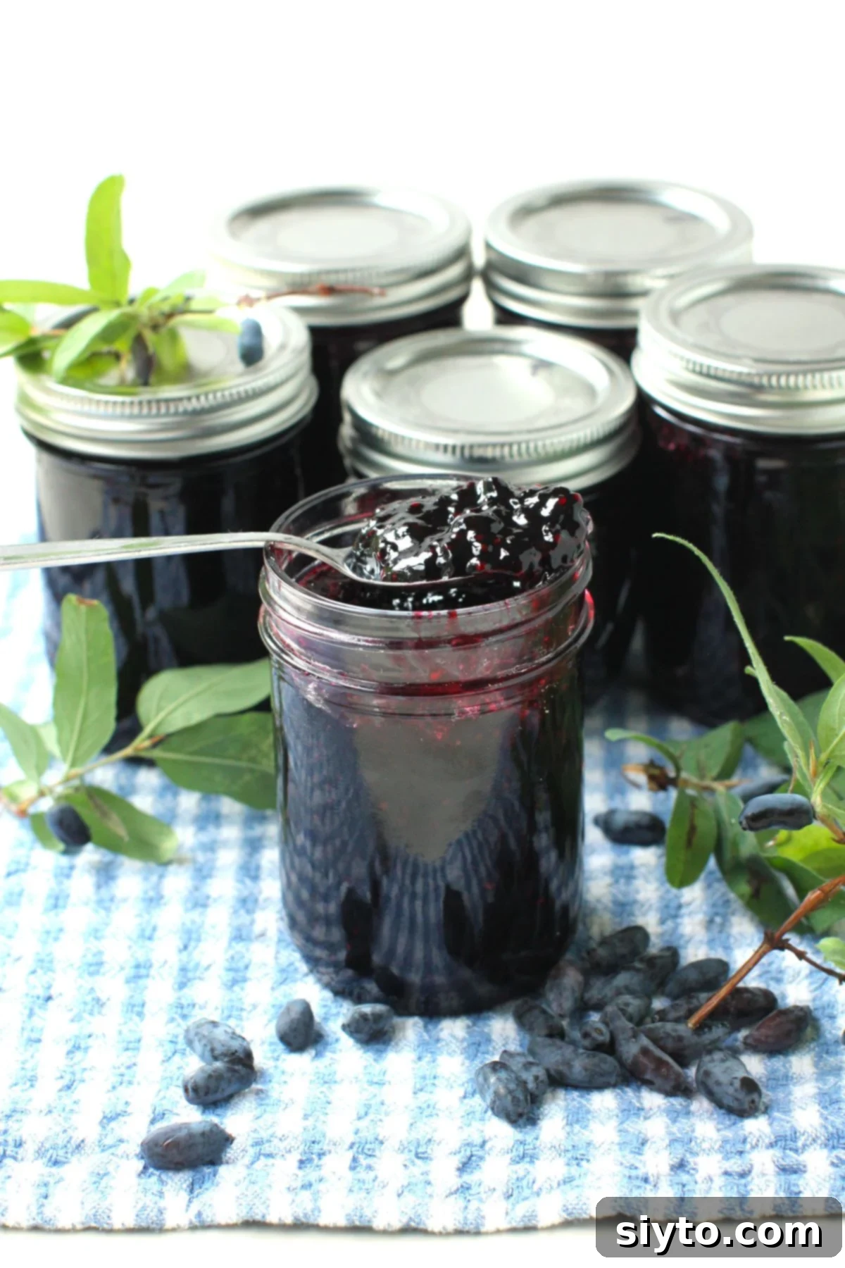 Homemade Haskap Honeyberry Jam Without Pectin 12 A jar of homemade haskap (honeyberry) jam, with a spoonful of jam propped on top. Fresh haskap berries are scattered on a towel, and more jars of jam are visible in the soft background.