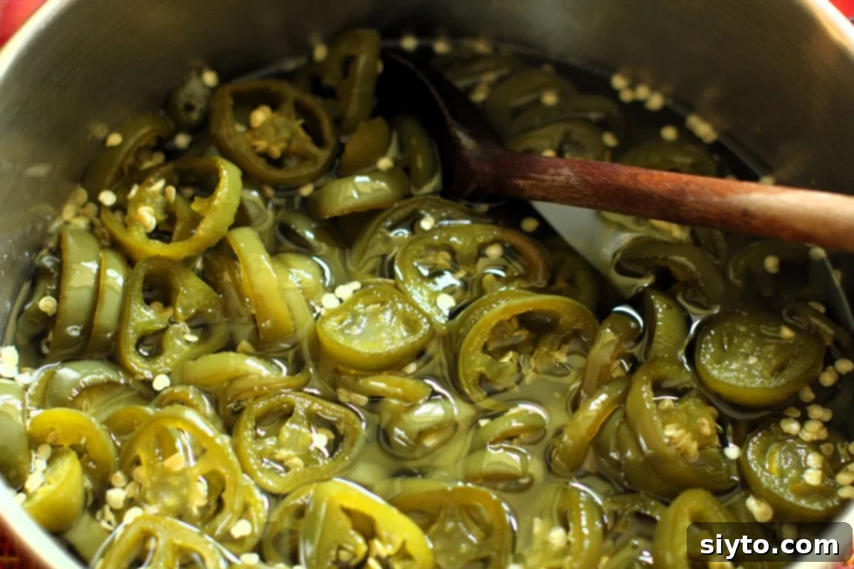 Sliced jalapeno peppers simmering in a sweet and tangy brine within a saucepan, turning a dull green color.