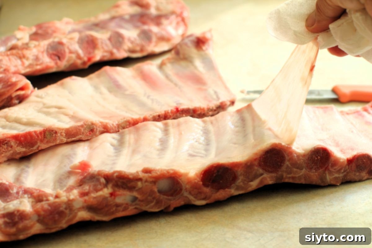 A close-up shot showing the process of carefully pulling the membrane off a rack of raw pork ribs, demonstrating an optional step for preparing ribs.