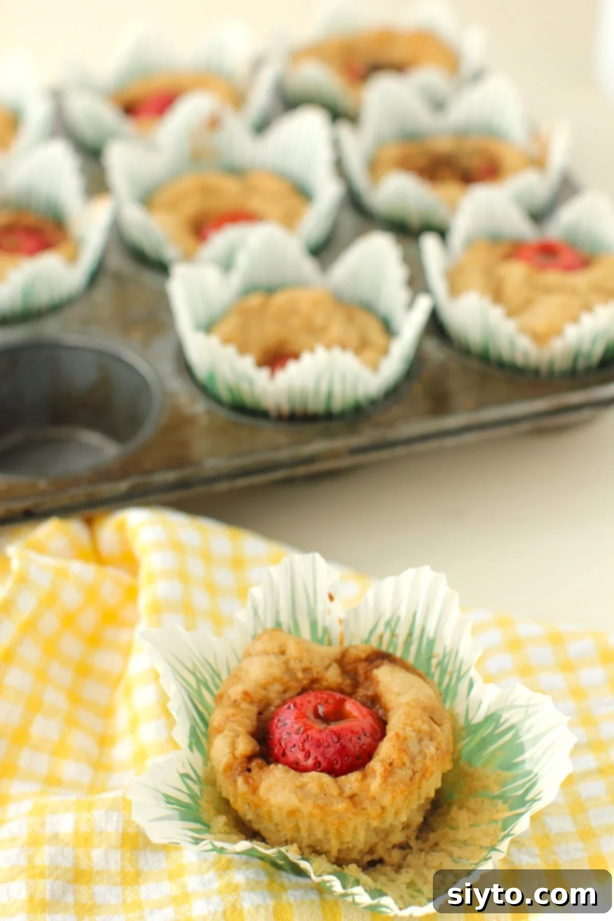 tray of baked muffins in the back, a muffin on the pulled paper liner in the front on top of a yellow-checked tea towel.