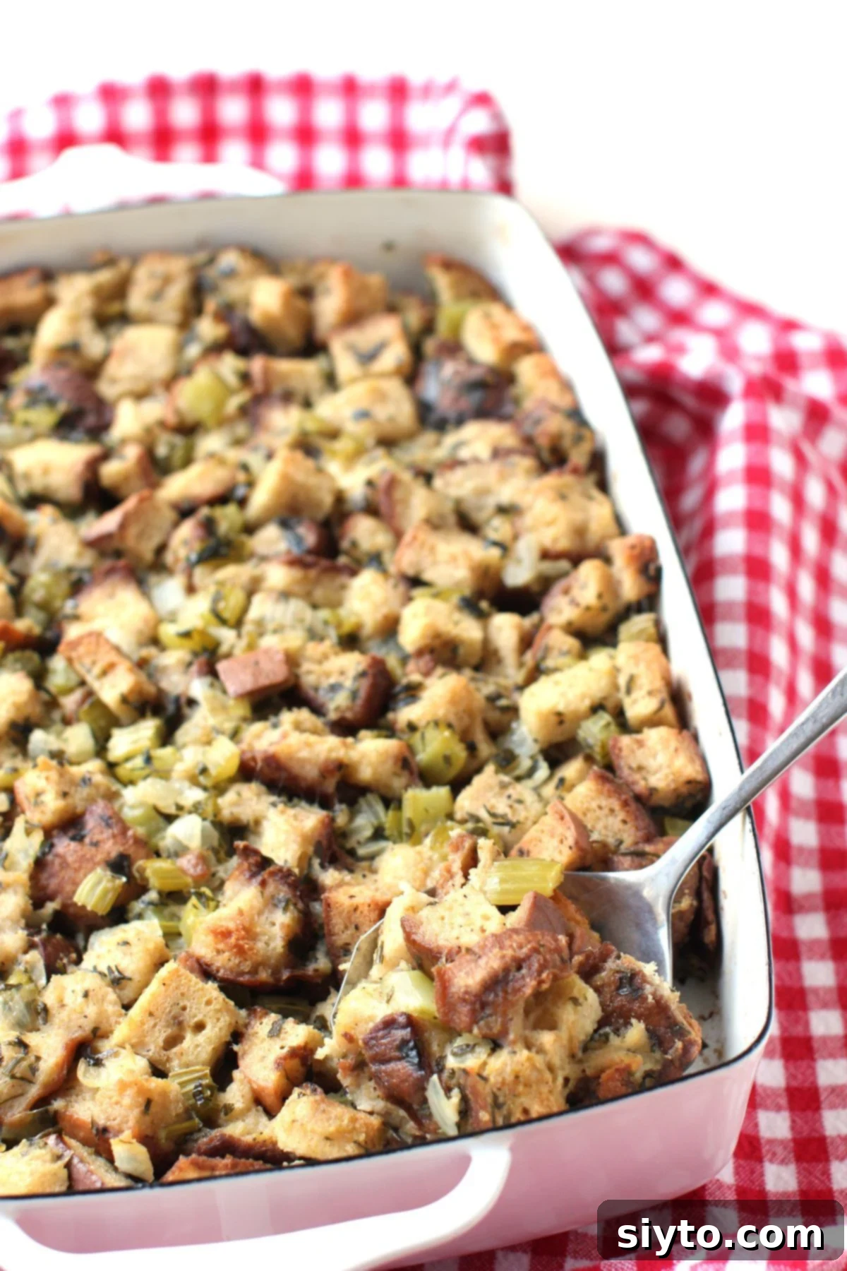 A spoonful of delicious gluten-free stuffing being scooped from a baking dish, set on a festive red and white checked tea towel, showcasing its golden-brown top and tender interior.