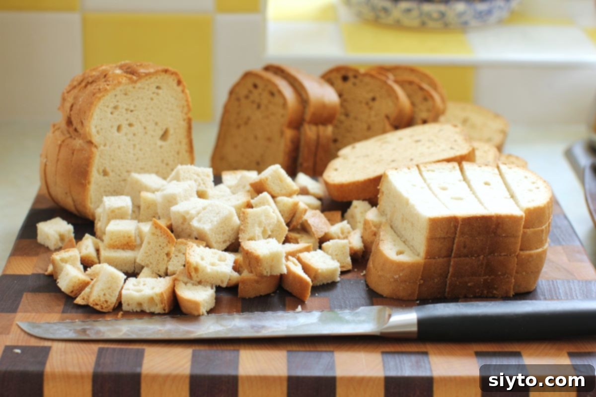 A cutting board with various stacks of white and whole-grain gluten-free bread slices, with some already cut into cubes, ready for stuffing preparation.