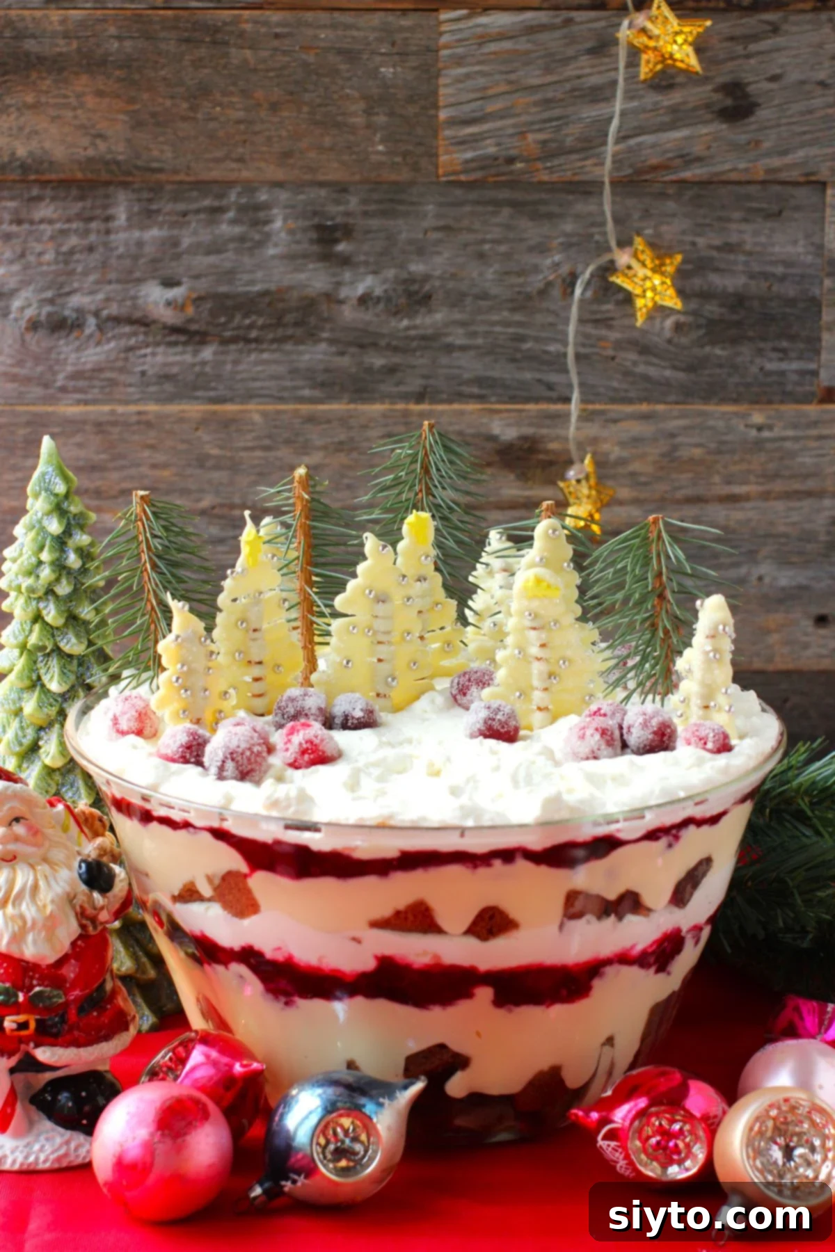 big bowl of Christmas trifle decorated with white chocolate trees, against a wood board background and glass ball ornaments in front.