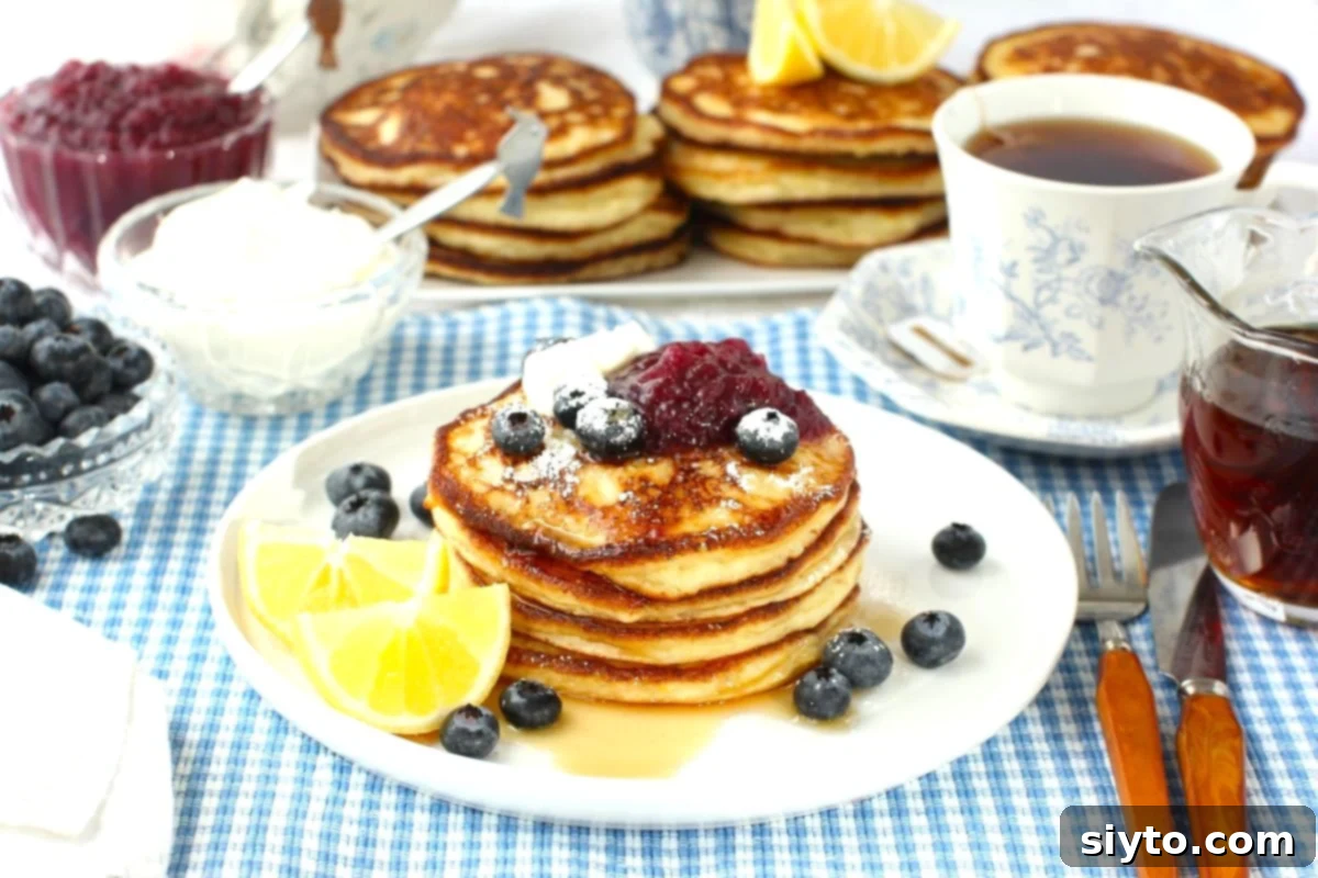 Fluffy Lemon Ricotta Pancakes 3 white plate with lemon ricotta pancakes in a stack, lemon wedges, blueberries, blueberry applesauce, tea in background and stacks of pancakes.