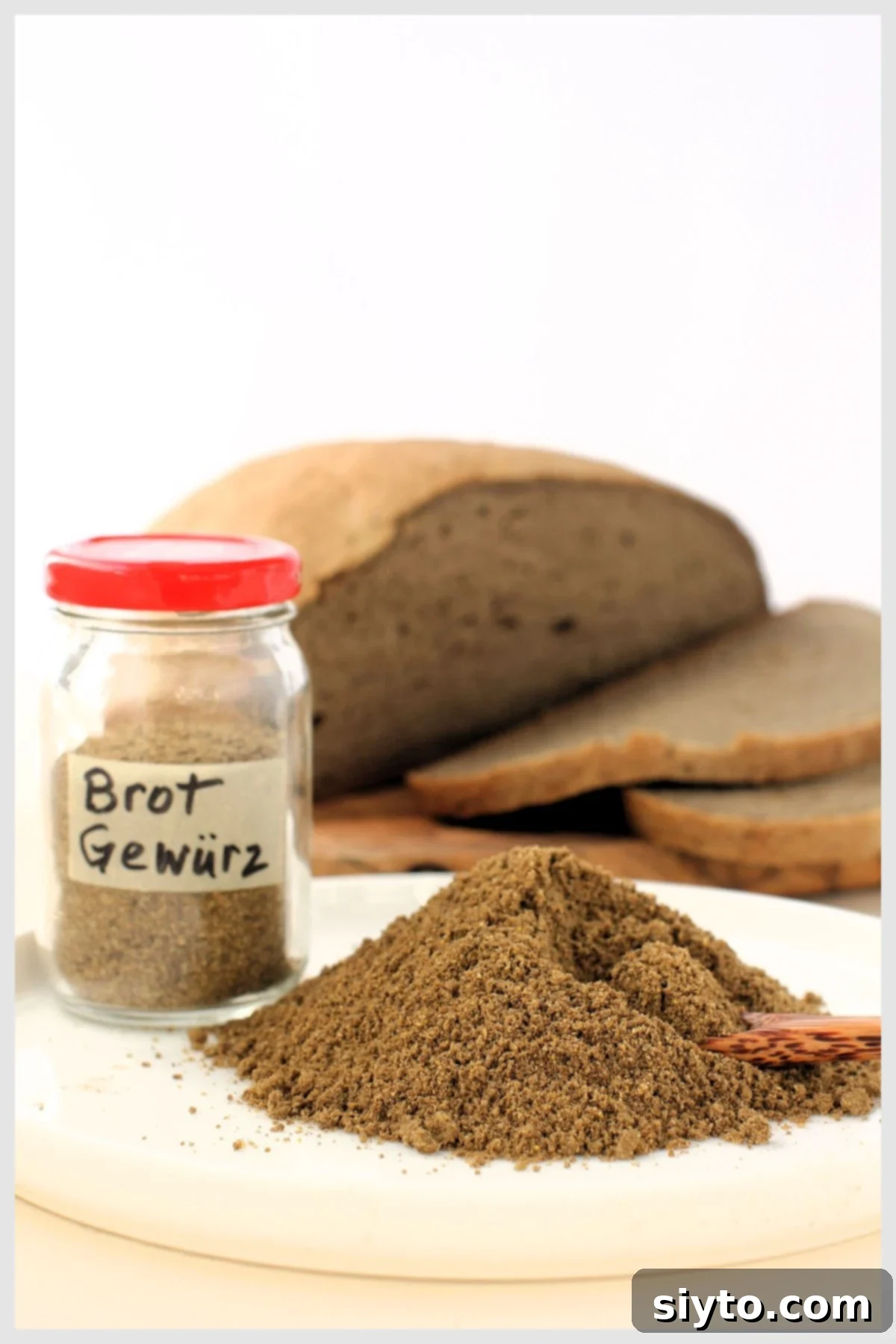 Jar of Brot Gewürz with red lid beside mound of bread spice, loaf of rye-style sliced bread behind.