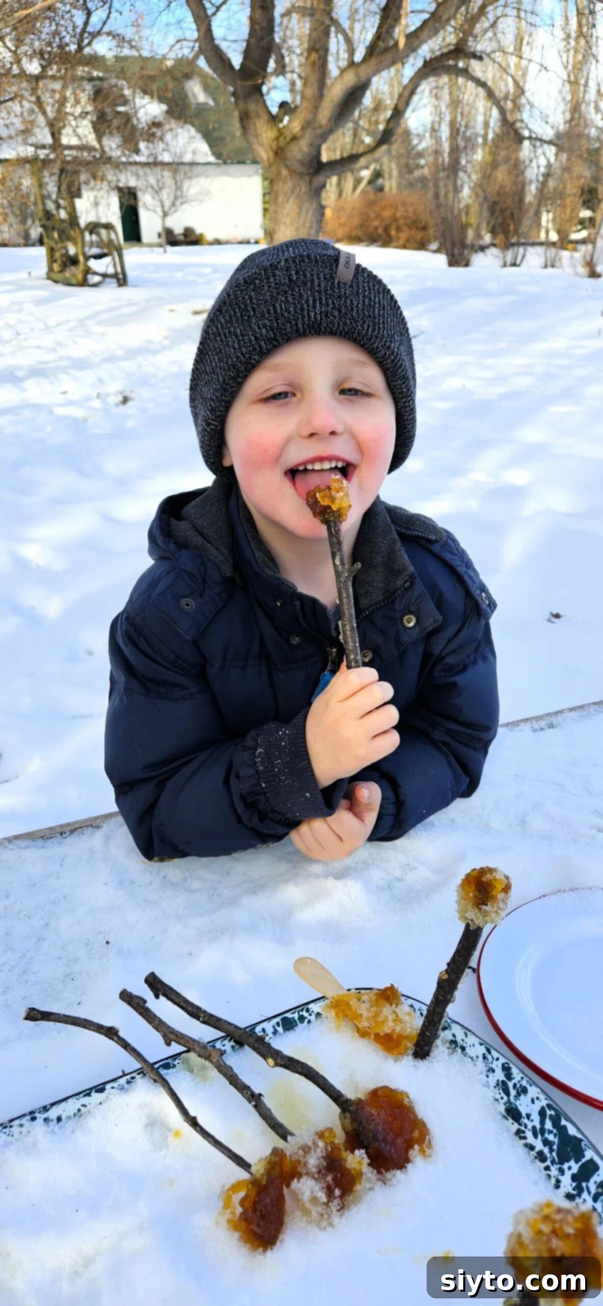 A young boy named Caleb happily eating maple syrup snow candy off a twig outdoors, his face beaming with a big smile.