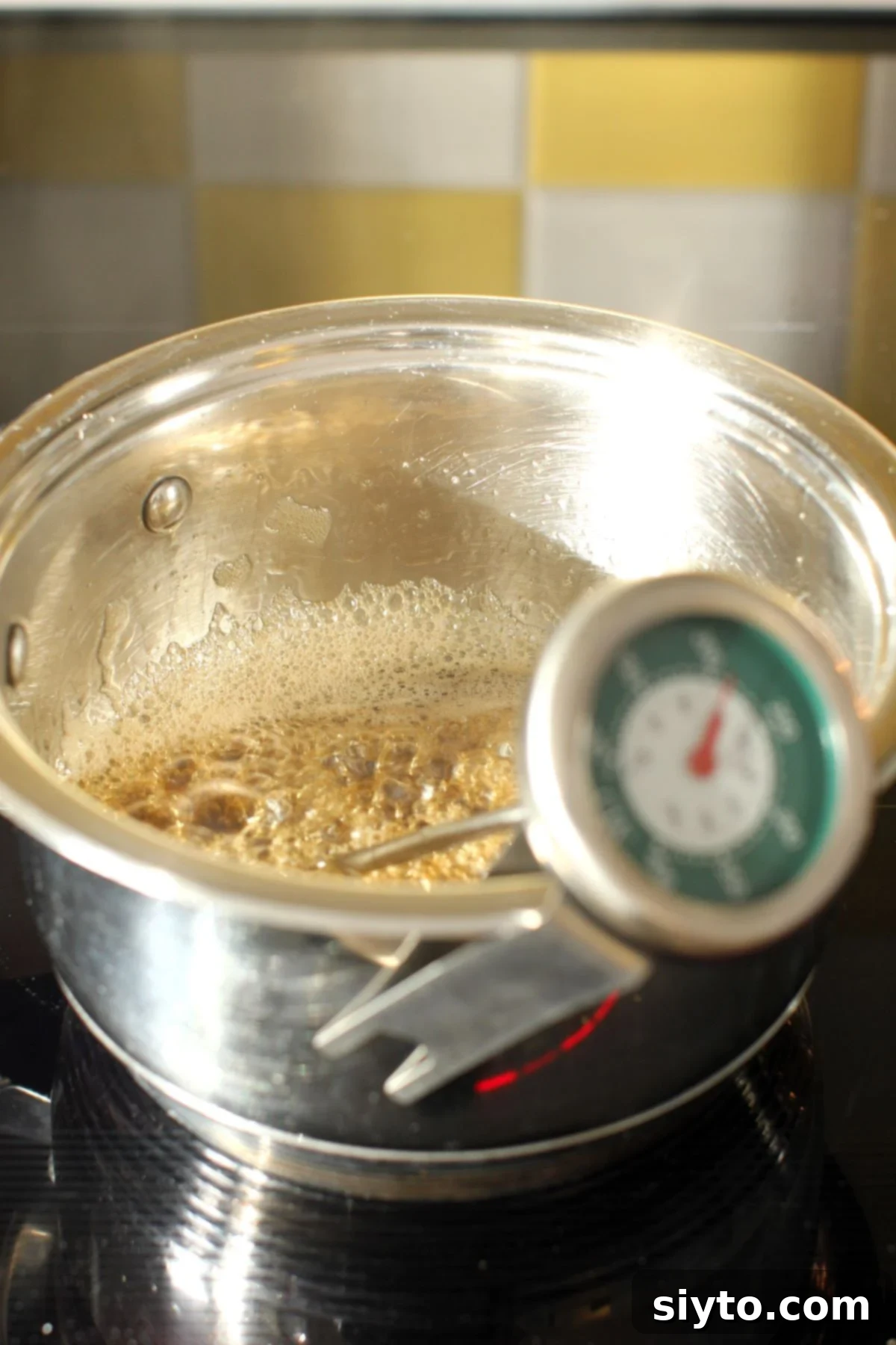 A saucepan of boiling maple syrup with a candy thermometer clipped to its side, sitting on a kitchen stove.