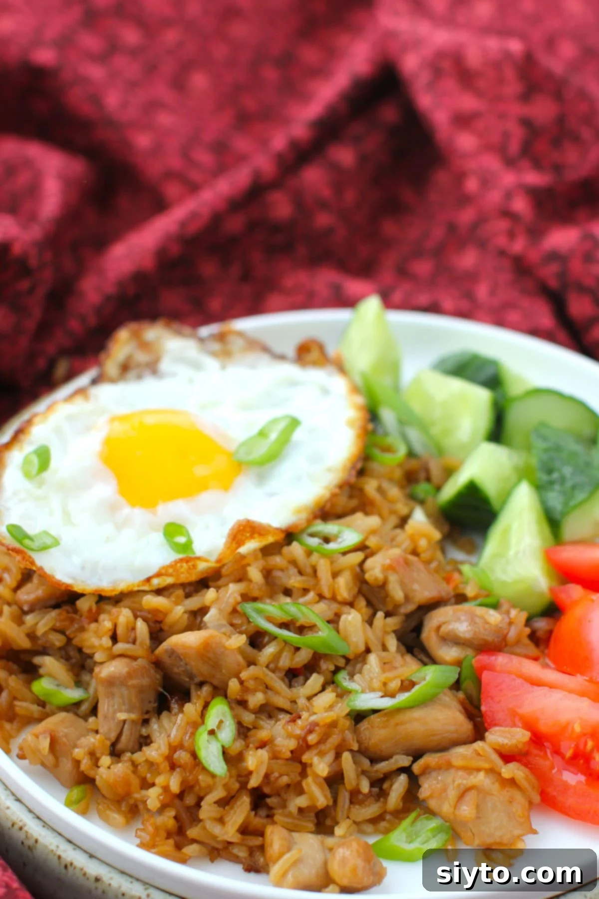 Indonesian Fried Rice Nasi Goreng 2 plate of Indonesian fried rice, with fried egg on top, tomatoes and cucumbers, against a burgundy cloth.