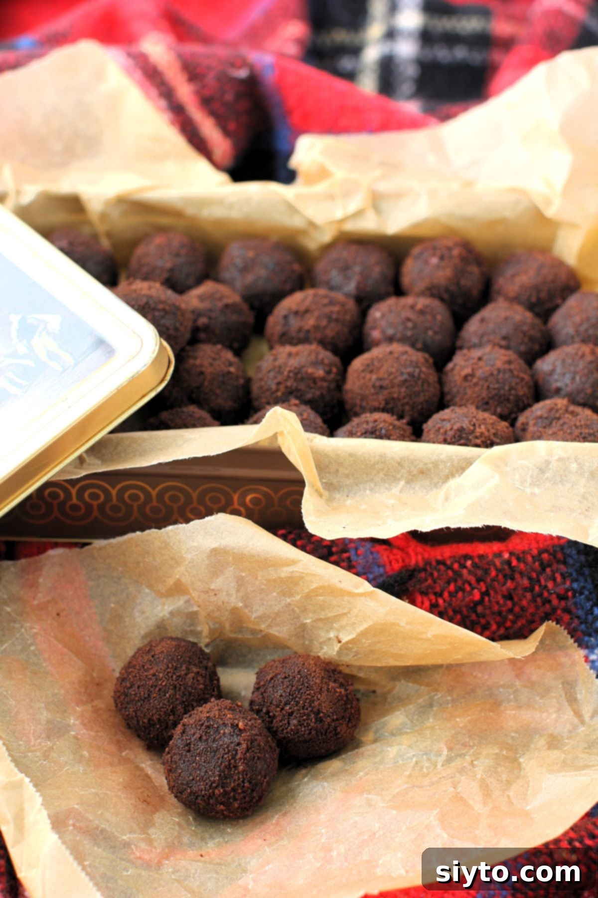 Sweet Bites Snowy Sleds: Lake Louise Adventures 2 3 glossy black licorice balls on crumpled parchment paper, with a tin full of more balls in the background, all resting on a vibrant red plaid blanket.