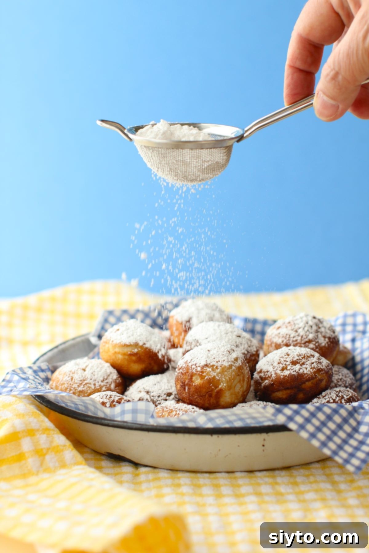 Shaking powdered sugar from a sieve onto a plate of Aebleskiver, resting on a charming yellow checked tea towel.