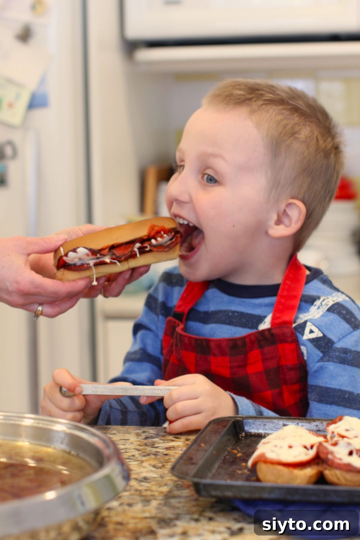 Caleb, a young boy, eagerly takes a bite of a pizza dog held out by an adult.