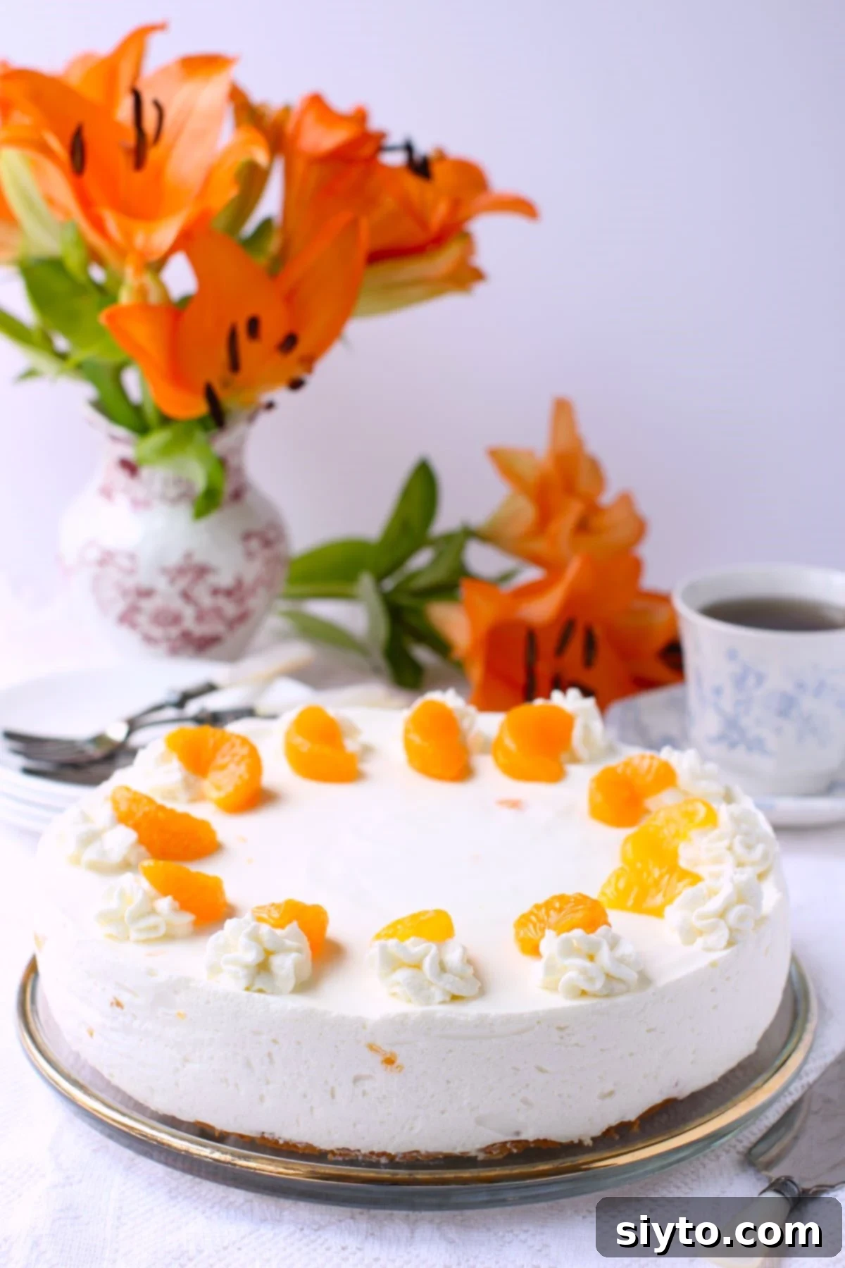 Luscious Mandarin Cream Torte 2 A beautifully decorated Mandarinen Sahnetorte, a German Mandarin Cream Torte, on a table. Behind it, a vase with vibrant orange lilies adds a touch of elegance, highlighting the torte's citrus theme and festive appeal. The torte features a light sponge base topped with a creamy, gelatin-set mousse filled with juicy mandarin orange segments, and garnished with more oranges and cream rosettes.