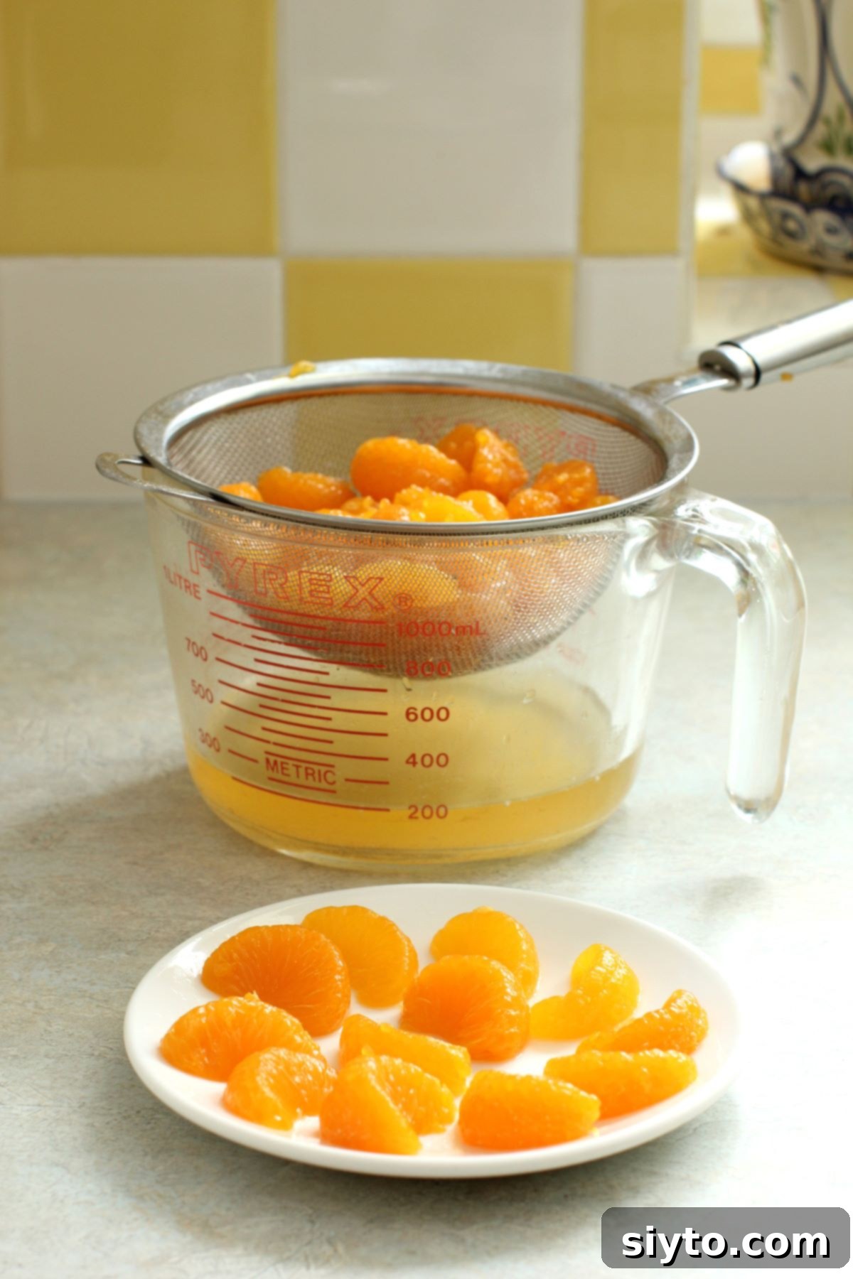 Luscious Mandarin Cream Torte 6 Mandarin orange sections are shown in a sieve, draining over a measuring cup to collect the juice. In front, a small plate holds a selection of perfectly preserved orange segments, set aside for garnish, illustrating the initial step of preparation for the torte.