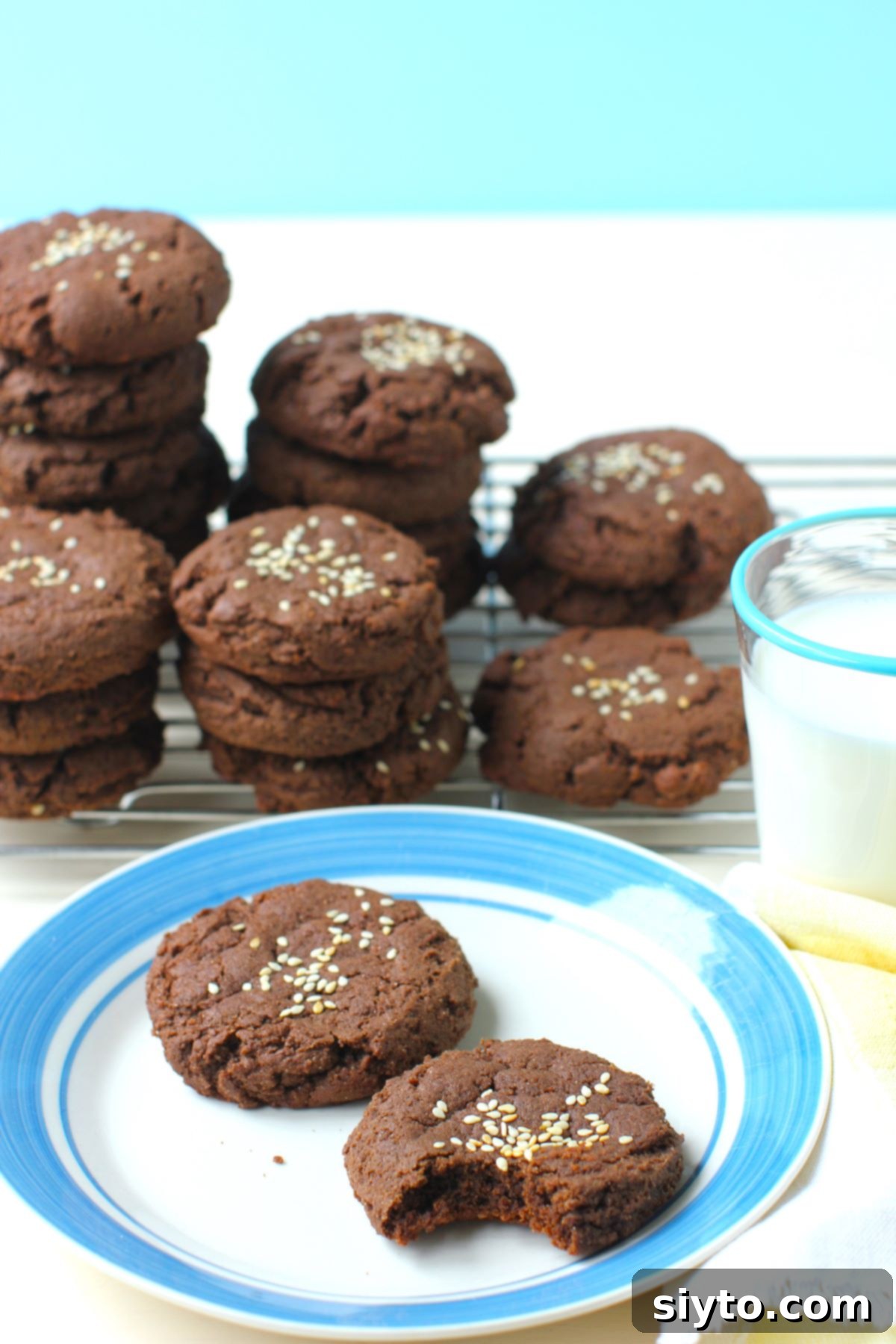 A plate featuring two rich chocolate tahini cookies, with more stacked in the background, showcasing their deliciously soft and fudgy texture.