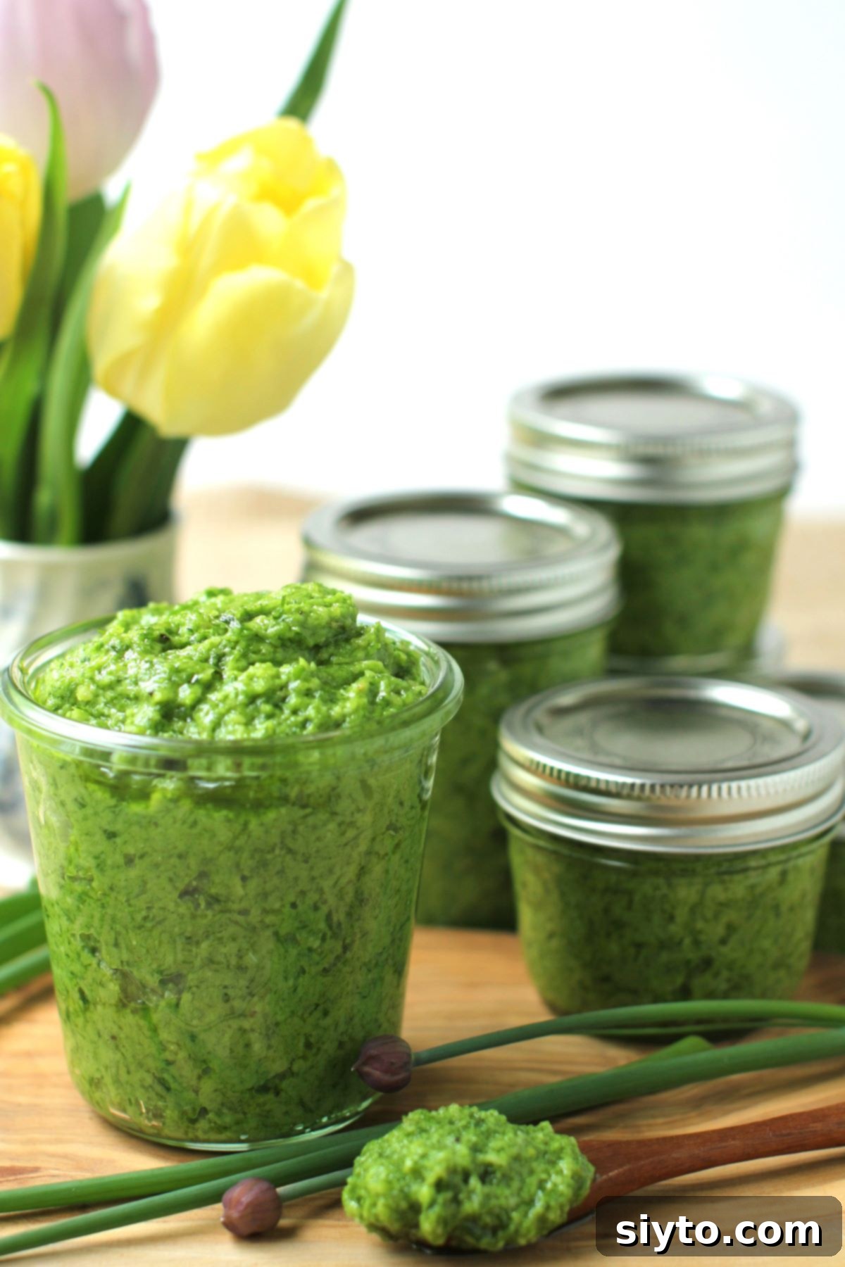 Chive Pesto: A Spring Garden Delight 2 A jar of freshly made chive pesto sits on a rustic wooden table, with other sealed jars of pesto blurred in the background, alongside a vase of cheerful spring tulips.