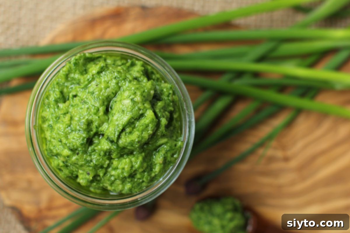 Chive Pesto: A Spring Garden Delight 13 A top-down view of a clear glass jar filled with vibrant green chive pesto, resting on a wooden cutting board, with fresh chive stems artfully arranged beside it.