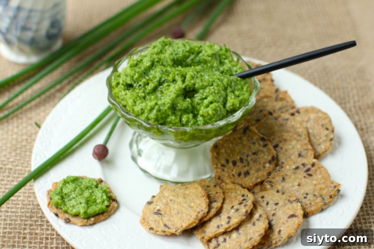 Chive Pesto: A Spring Garden Delight 14 A small white bowl filled with vibrant green chive pesto, surrounded by a selection of crunchy crackers, arranged artfully on a white serving plate.