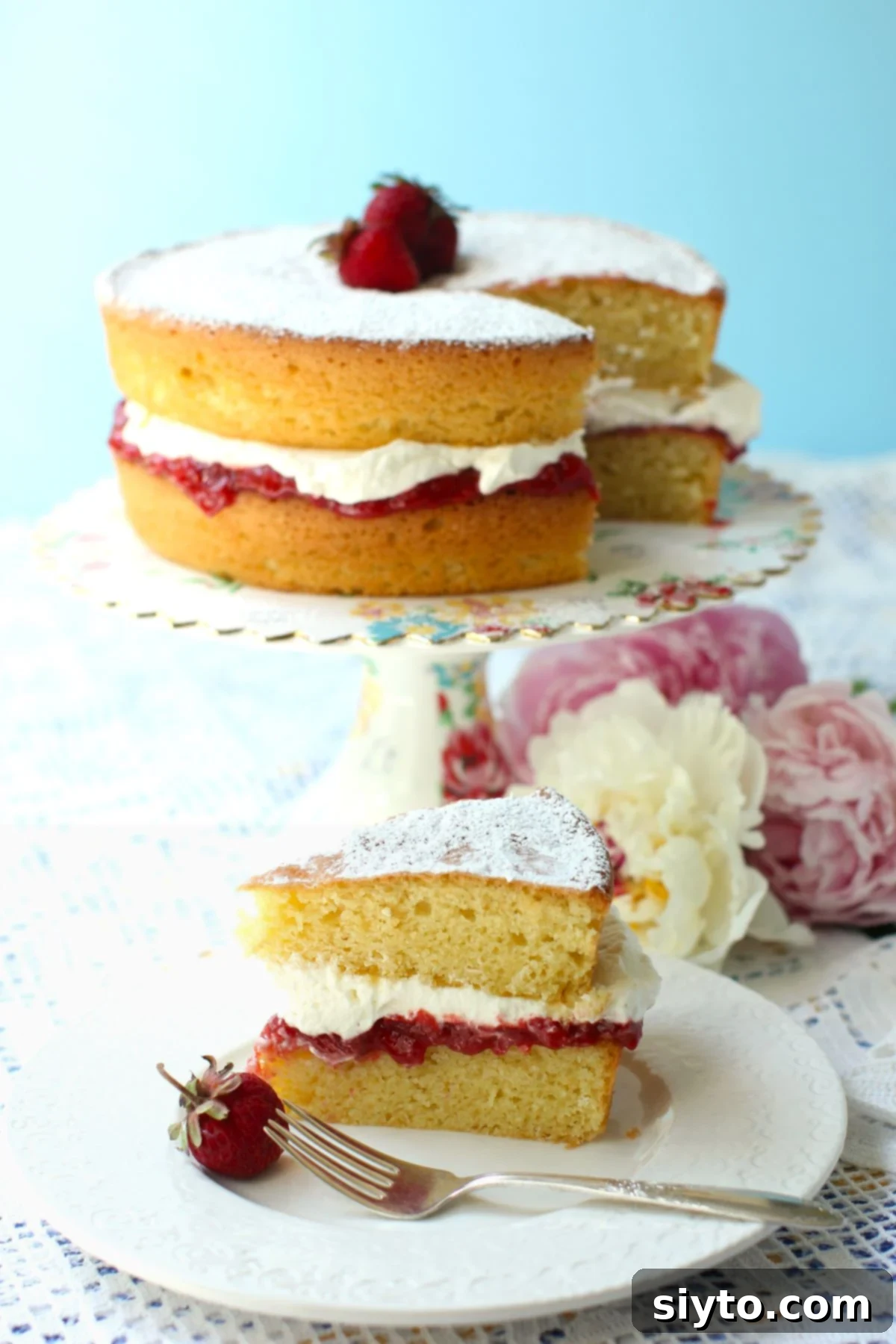 A beautifully sliced Gluten-Free Victoria Sponge Cake, showcasing its moist layers filled with jam and cream, presented on a elegant cake stand with a piece on a plate in front.