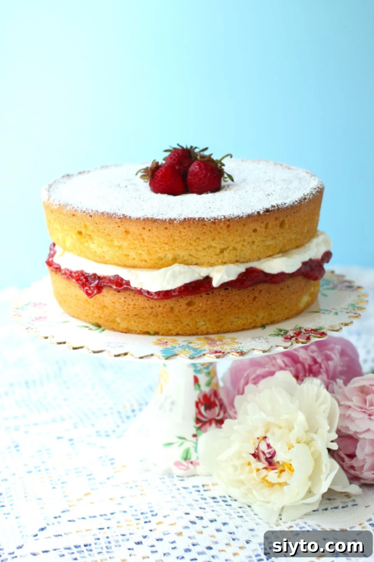 A beautiful Gluten-Free Victoria Sponge cake, garnished with fresh peonies lying beside it, presented on an elegant cake stand.