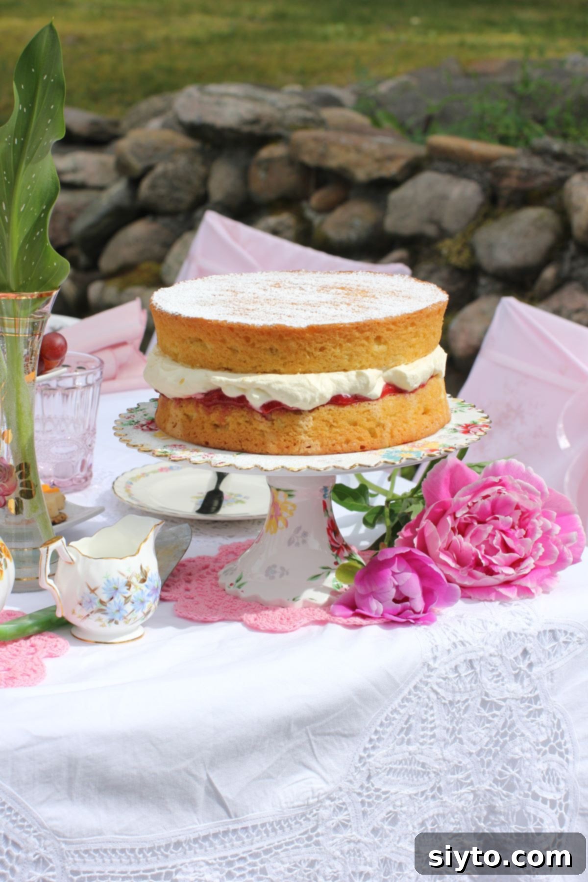 The elegant Gluten-Free Victoria Sponge cake graces a cake stand, beautifully displayed on a lace-covered picnic table, ready to be enjoyed.