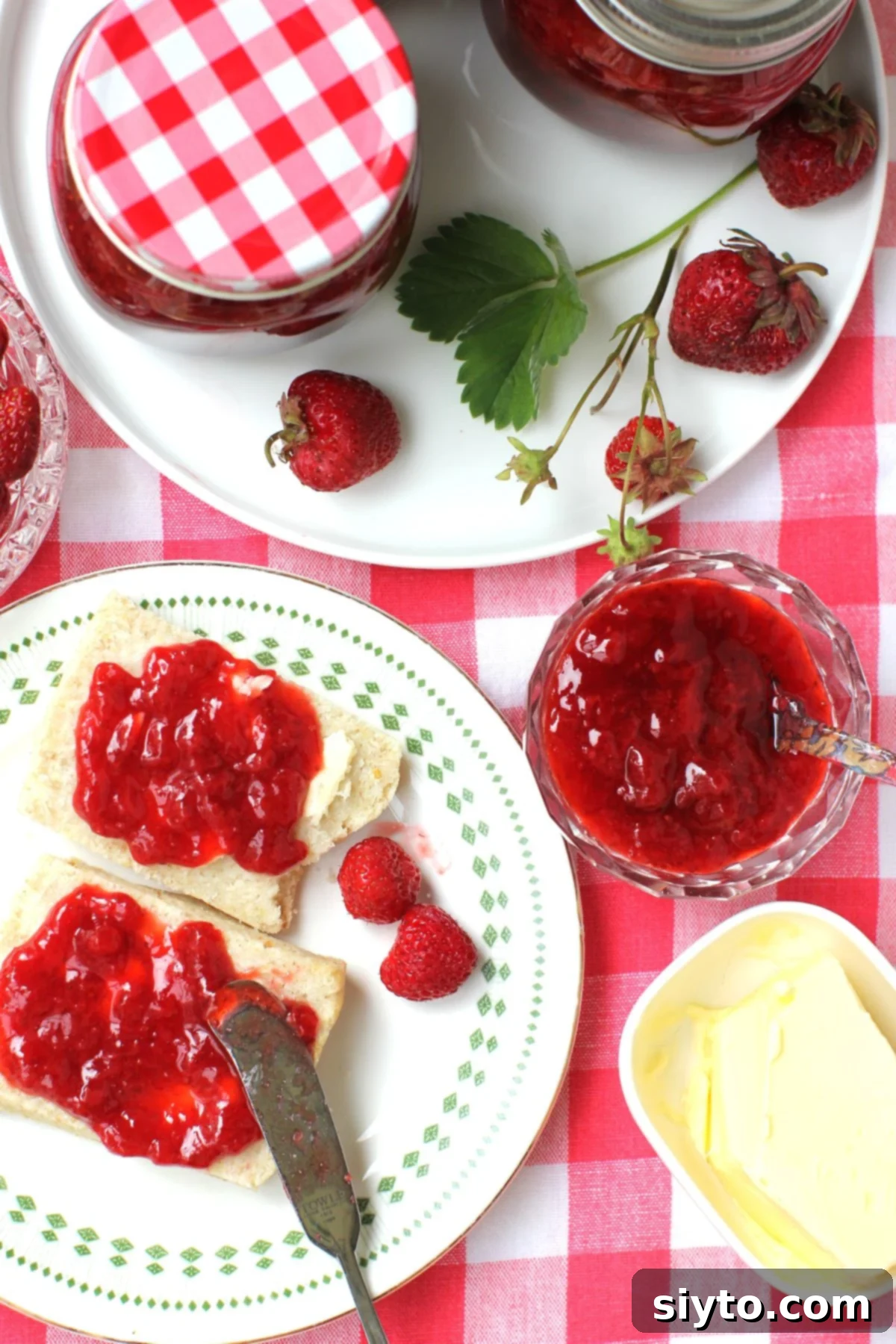Reduced Sugar Strawberry Jam 2 Looking down at the table with a red-checked tablecloth, plate of two biscuit halves spread with low sugar strawberry jam, and butter.
