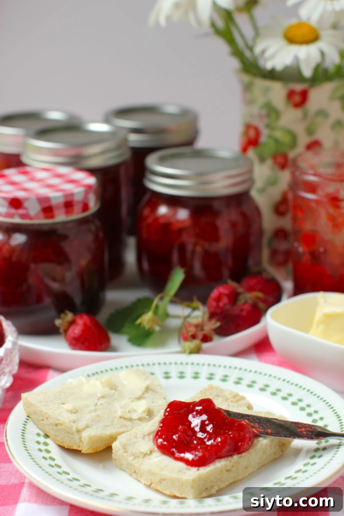 Reduced Sugar Strawberry Jam 9 Biscuit spread with low sugar strawberry jam on a plate, with jars of jam in the background.