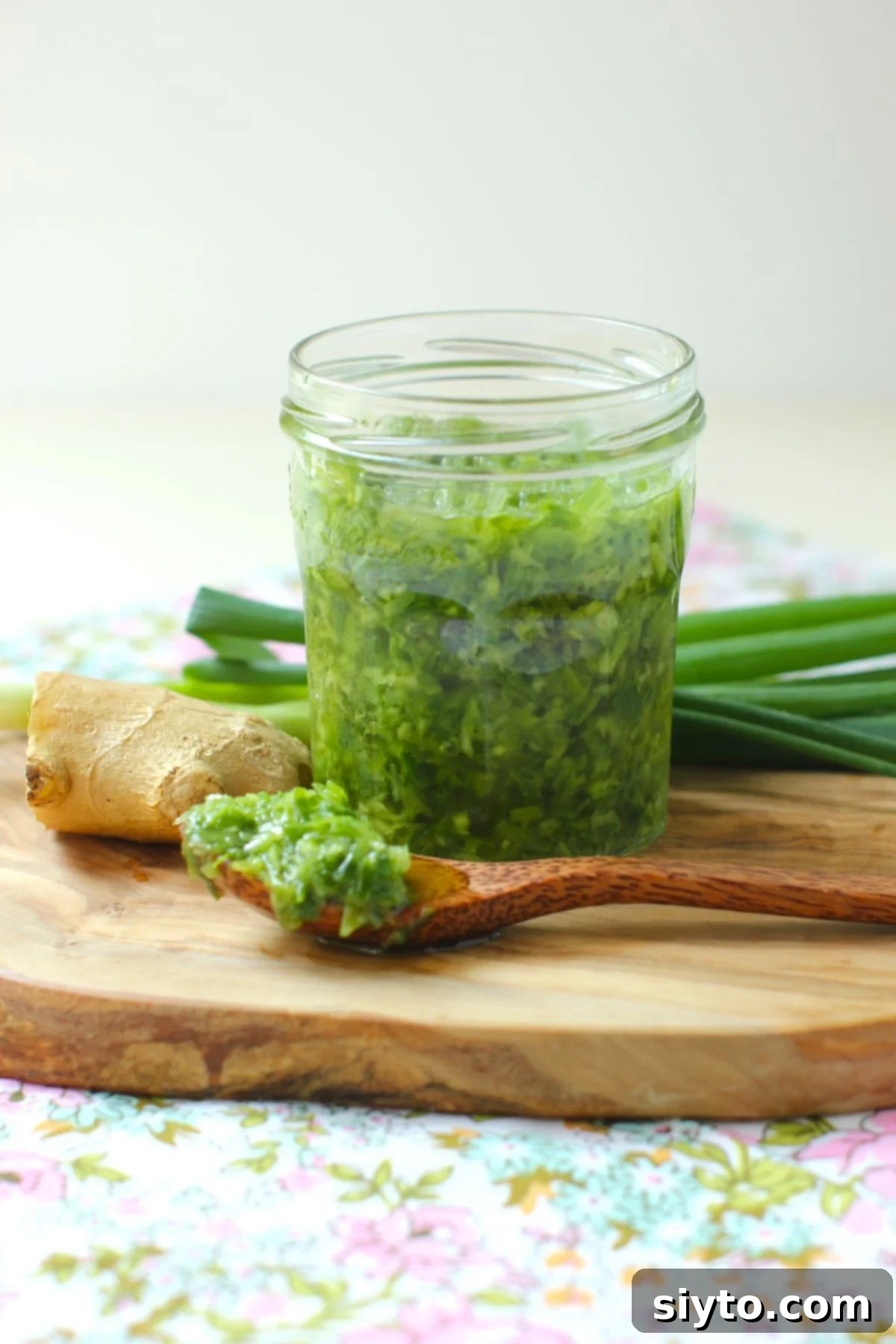 A jar of homemade ginger scallion sauce on a wooden cutting board, with a spoonful of sauce in front, and fresh ginger and scallions arranged behind it.