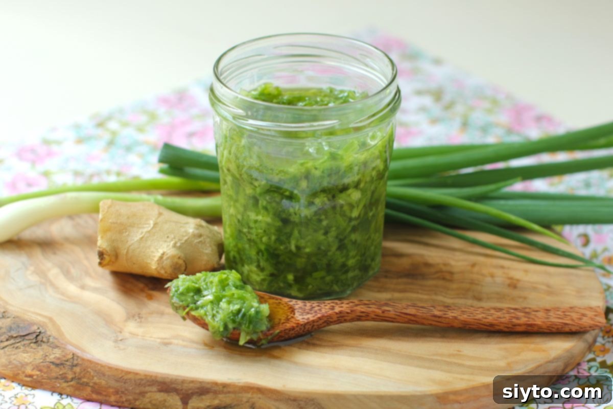 A glass jar filled with vibrant ginger scallion sauce sits on a wooden cutting board. Fresh green onions and a knob of ginger are in the background, with a spoon full of the golden-green sauce resting in front.
