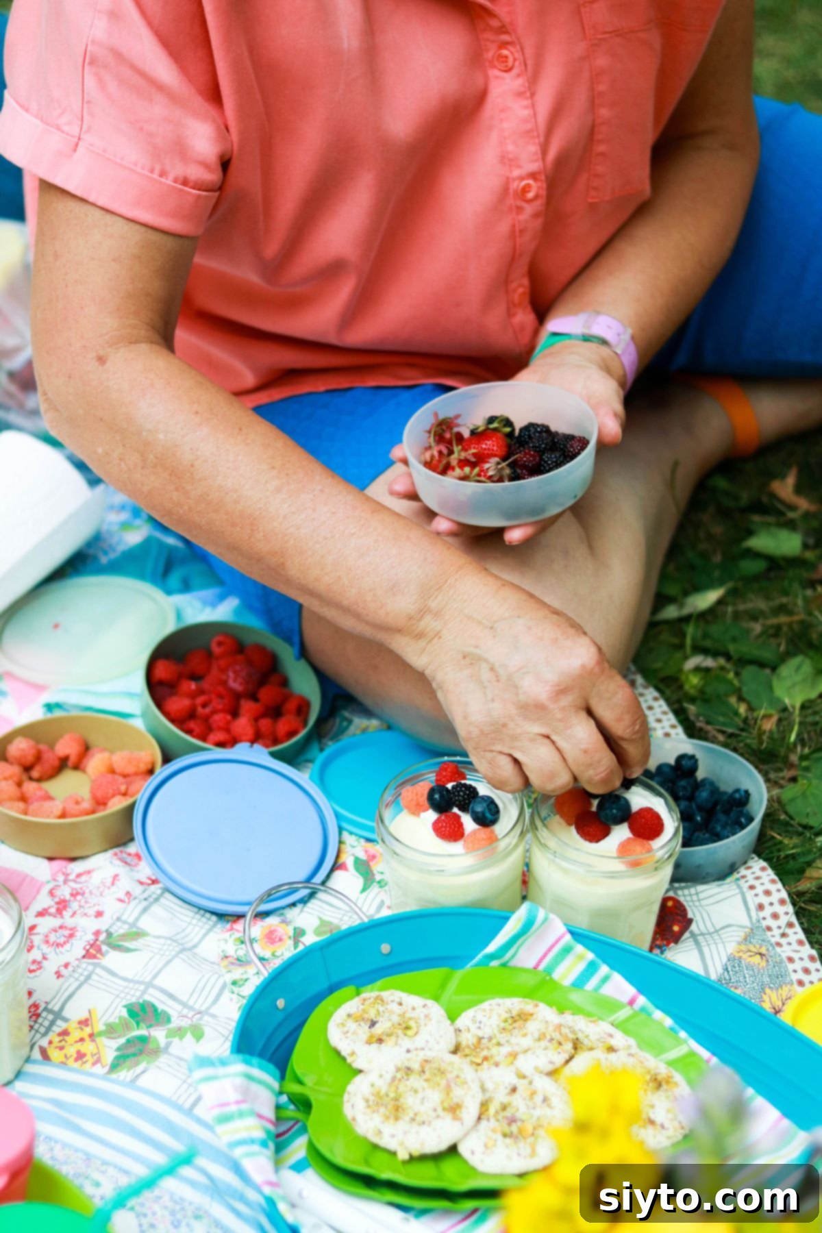 Sitting on a picnic blanket, a person adds fresh garden berries to individual jars of lemon mousse, enhancing the dessert for outdoor enjoyment.