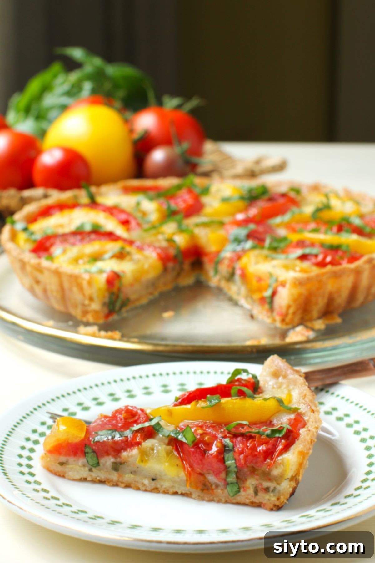Rustic Tomato Tart 7 A perfectly baked slice of tomato tart on a white and green plate, with the rest of the tart and fresh tomatoes blurred in the background.