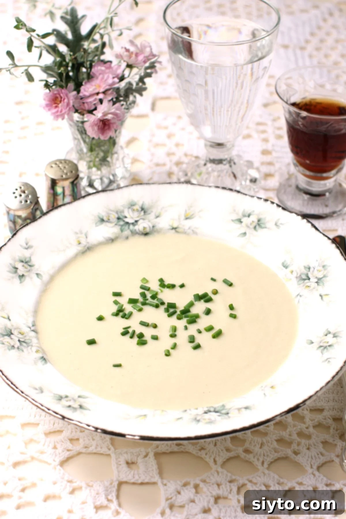 A rimmed china bowl with cream of puffball soup garnished with chopped chives, on a lace tablecloth, with small vase of pink flowers and glass of sherry.