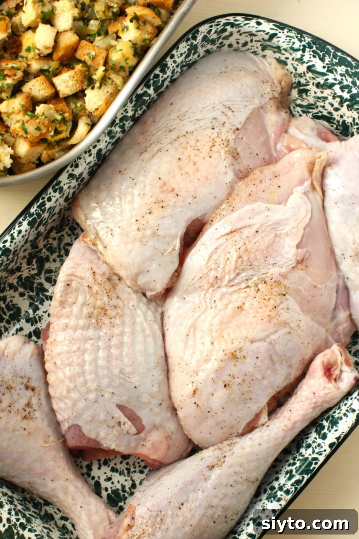 A roasting pan filled with savory stuffing sits beside a tray of seasoned turkey pieces (drumsticks, thighs, and breast cuts) rubbed with oil and sprinkled with salt and pepper, ready to be placed on top of the stuffing.