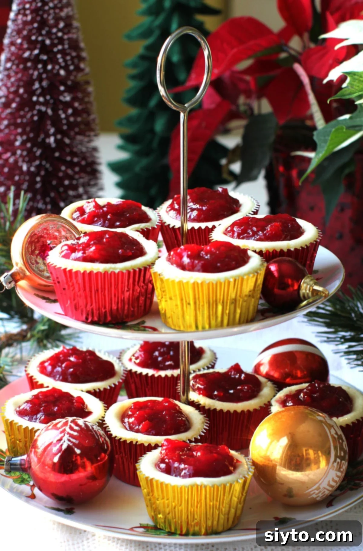Two-tiered serving plate showcasing beautifully arranged cheesecake cupcakes in festive red and gold foil liners, adorned with glass Christmas tree ornaments.