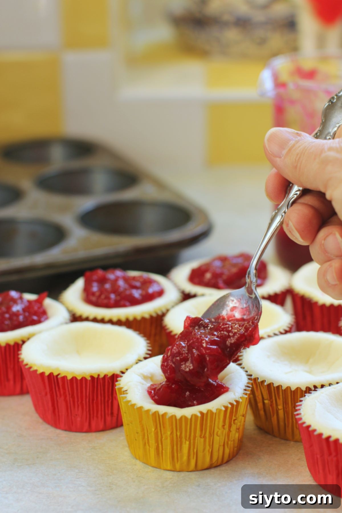A hand elegantly spooning vibrant cranberry orange topping onto a freshly baked cheesecake cupcake, highlighting the final delicious step.