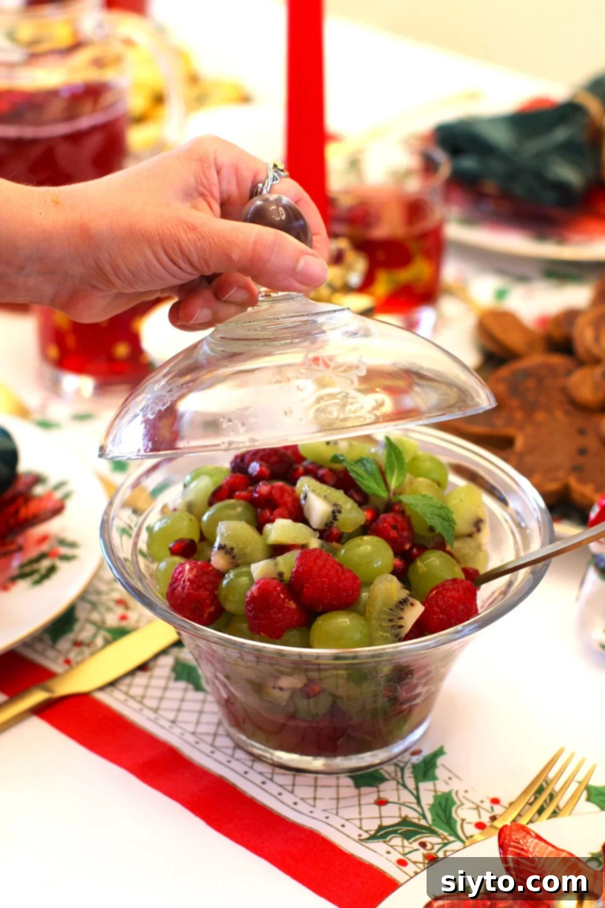 A glass lid being lifted from a beautiful glass bowl filled with a colorful holiday fruit salad featuring green grapes and kiwis, and red raspberries and pomegranate seeds.