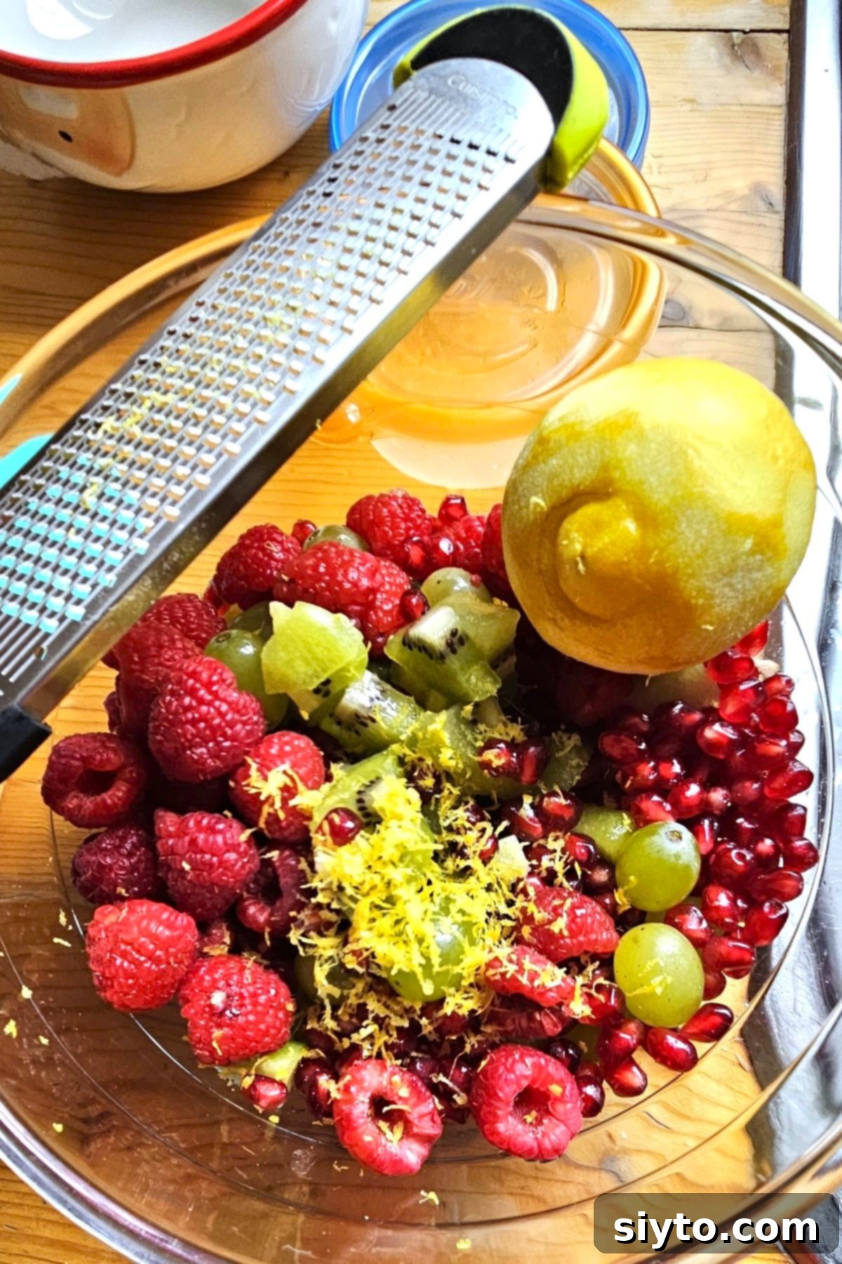A vibrant assortment of fresh fruits, including green grapes, kiwis, red raspberries, and a pomegranate, artfully arranged in a bowl, with a zested lemon and a rasp nearby for making the holiday fruit salad.