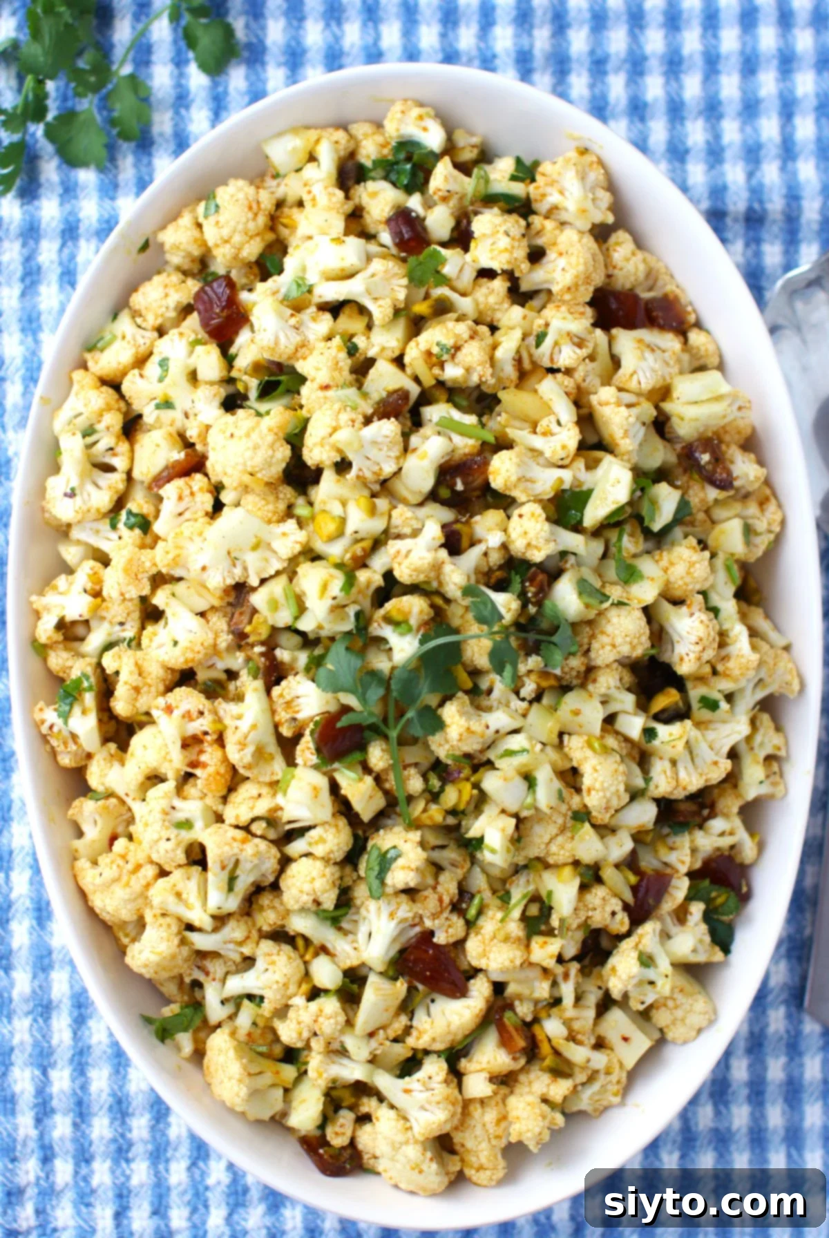 Looking down on an oval white bowl of cauliflower salad, on a blue and white checked tea towel.