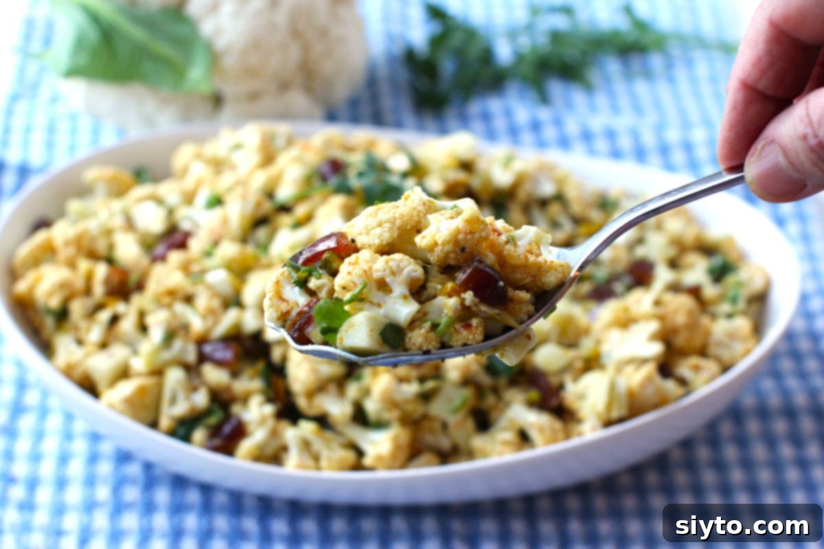 Close-up of a serving spoon full of cauliflower salad held in hand, with blurry bowl of salad behind.