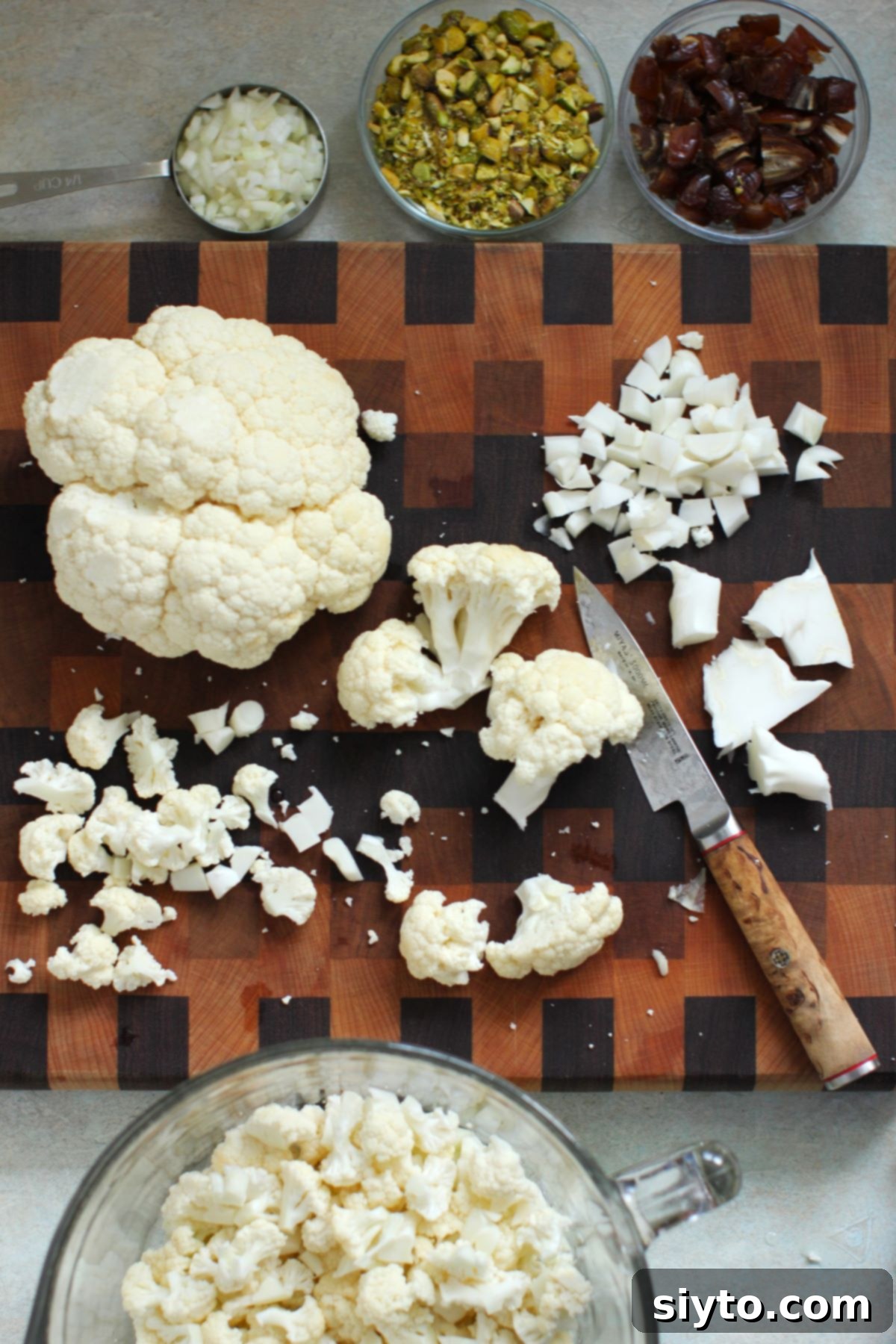 Cauliflower cut into florets and small dice on a checkered wooden cutting board, with bowls of chopped onion, pistachios, and dates beside it.